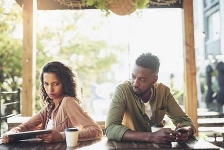 A man and woman sit at a table, holding a tablet and a phone. They are angled away from each other and look upset.